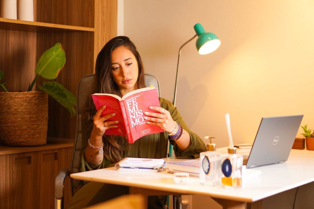 Bruna Biscalchini sentada em sua mesa, lendo um livro com capa cor magenta. Ao fundo, uma estante de madeira com livros e decoração minimalista. Sobre a mesa, um laptop e um abajur atrás com iluminação direcional.